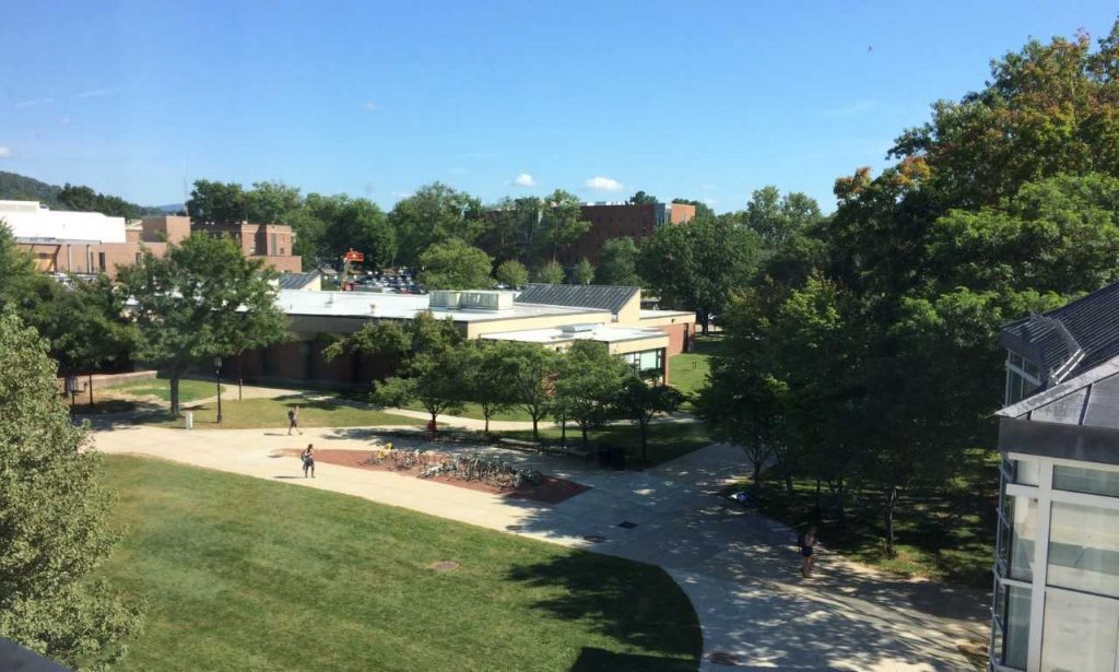 A photo taken from up high, looking down on a pathway where students are walking, a green lawn, a large bike rack, a one-story brick building, and deep blue sky with one fluffy white cloud in the center. 